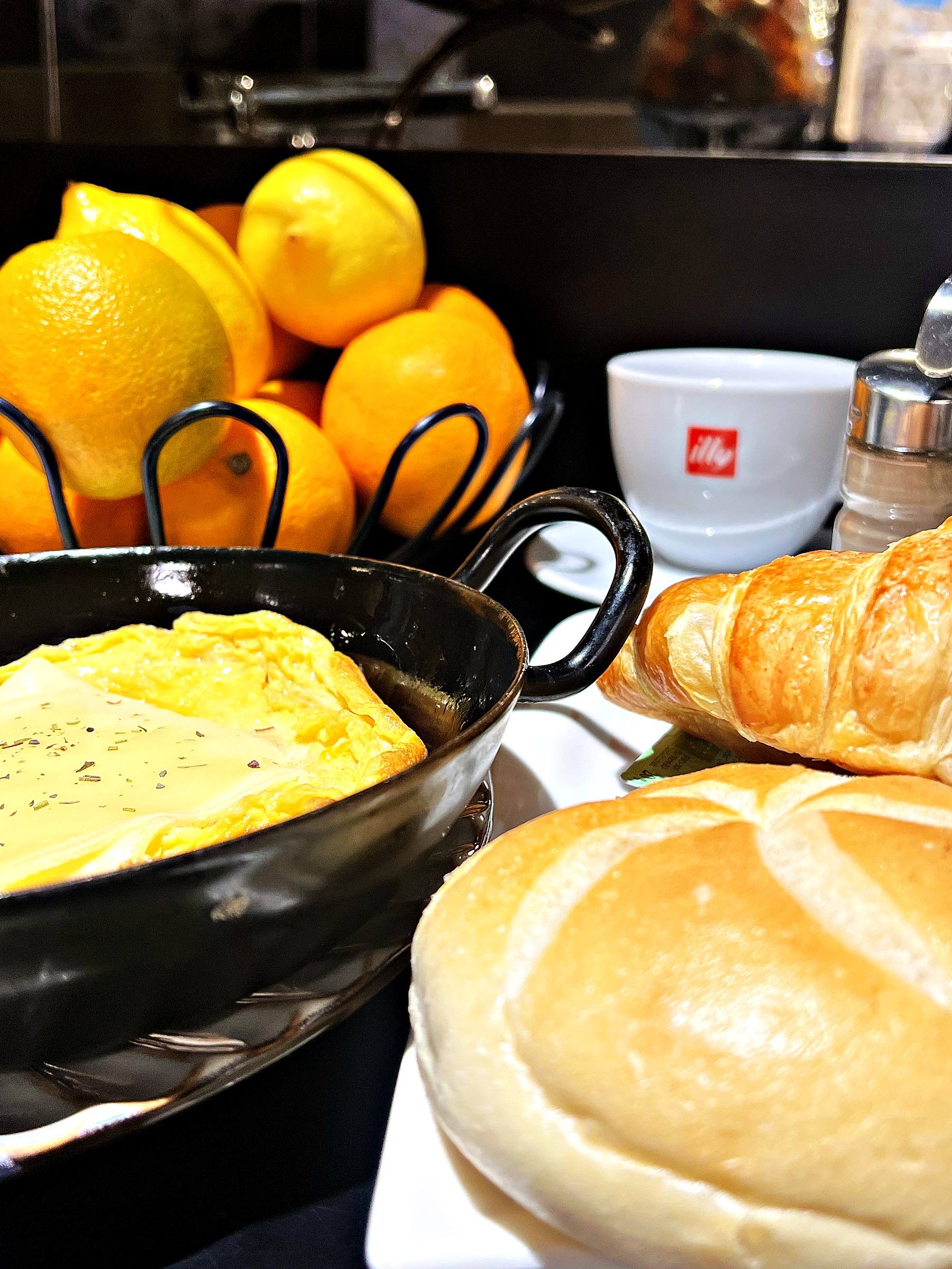 Frühstück mit Eierspeis in der Pfanne, Croissant, Semmel und einer illy-Tasse, dahinter Orangen