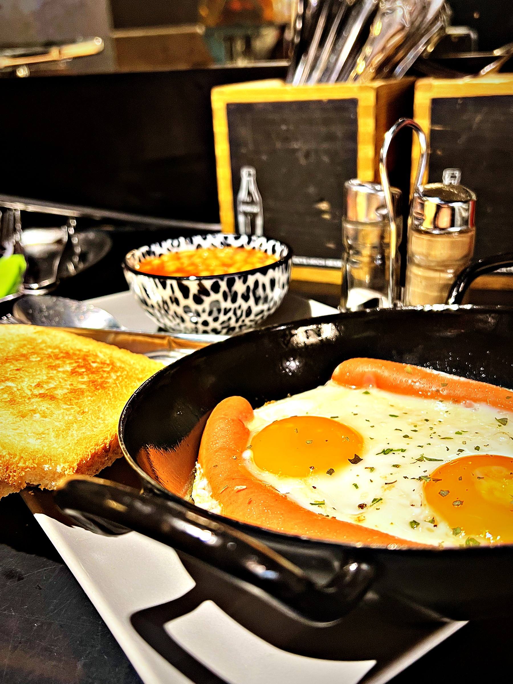 English Breakfast: zwei Spiegeleier und Würstel in der Pfanne, daneben Toast und Bohnen